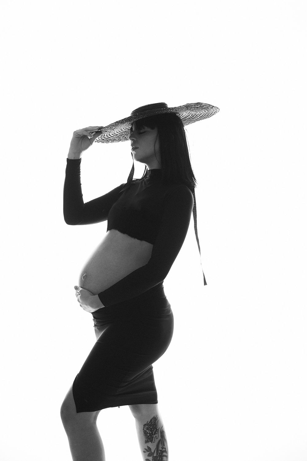 Dramatic black and white maternity portrait with wide-brimmed hat creating artistic silhouette.