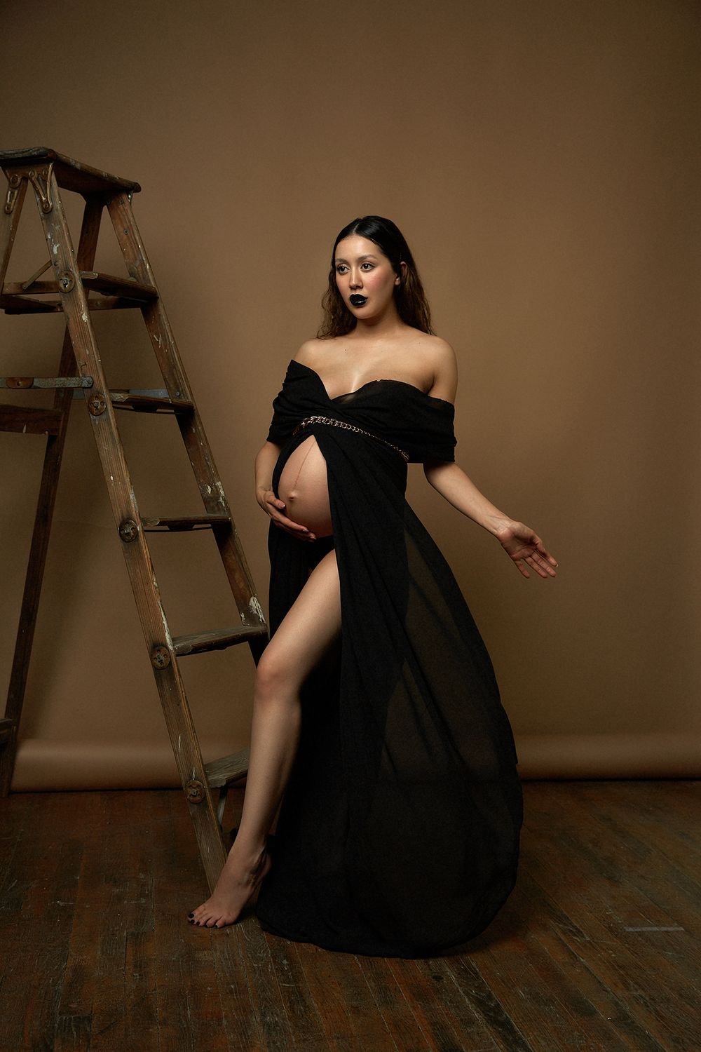 Dramatic maternity portrait in black off-shoulder gown beside vintage wooden ladder.