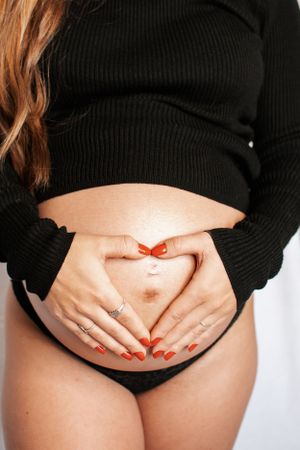 Close-up of pregnant belly with hands making heart shape and red nail polish.