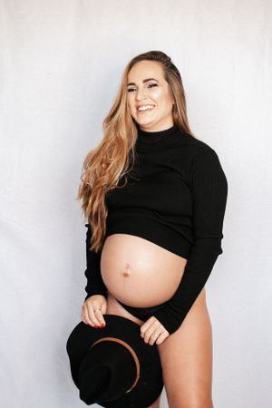 Maternity photo featuring woman in black turtleneck crop top holding brown hat.