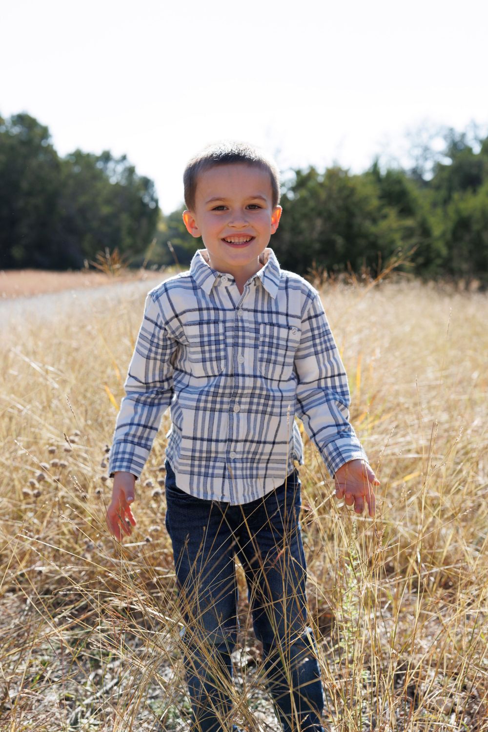 A young boy in a plaid shirt smiles while standing in a sunlit field of tall grass.