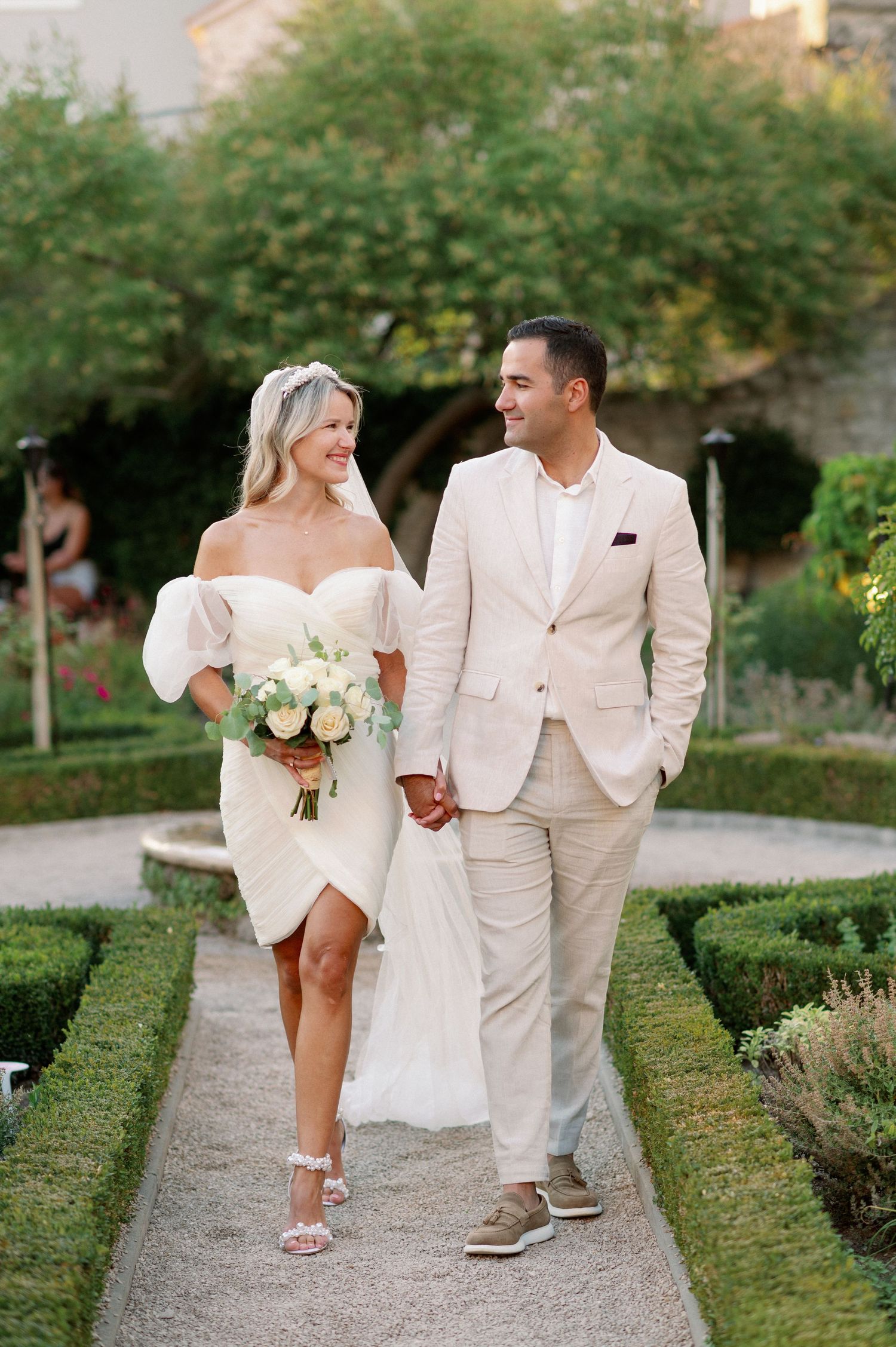 A couple walks hand in hand through a garden path, wearing elegant cream-colored wedding attire and holding a white bouquet.