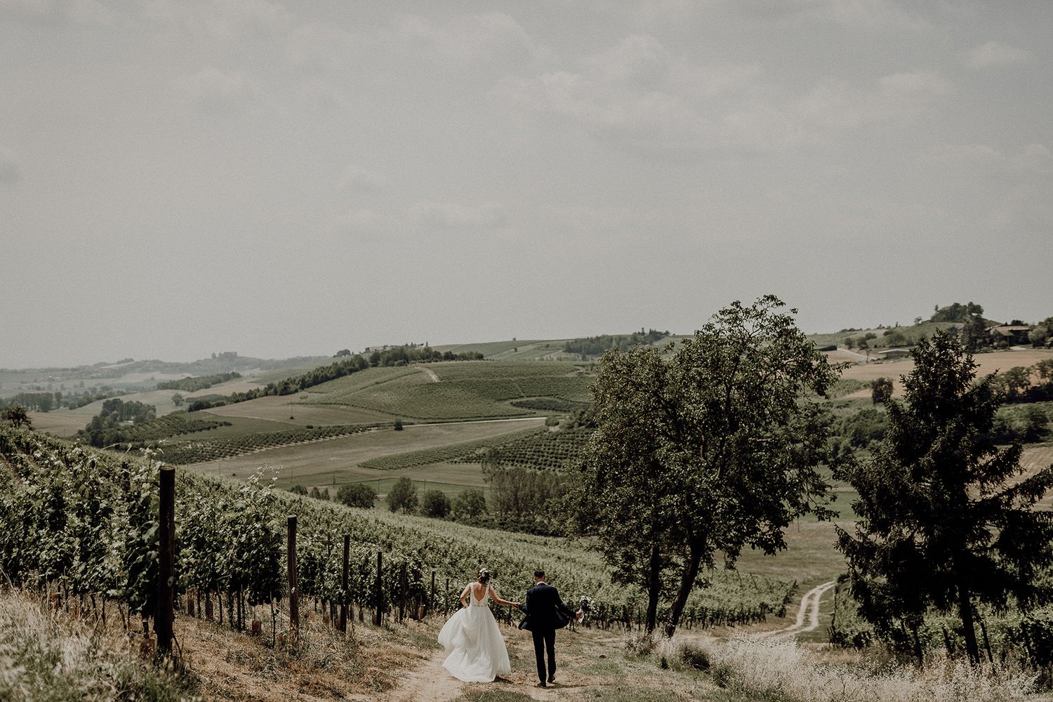 Bride and groom walk hand in hand along a vineyard path with sweeping Tuscan countryside views behind them.