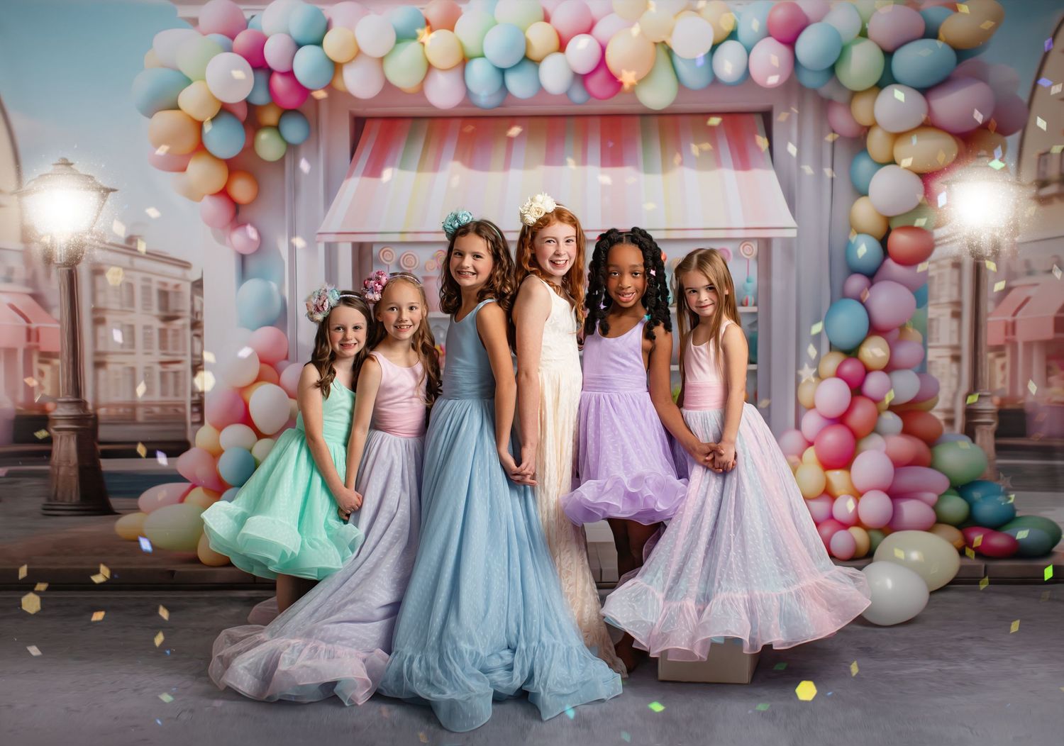 Group in pastel dresses posing against candy-colored balloon arch and storefront backdrop.