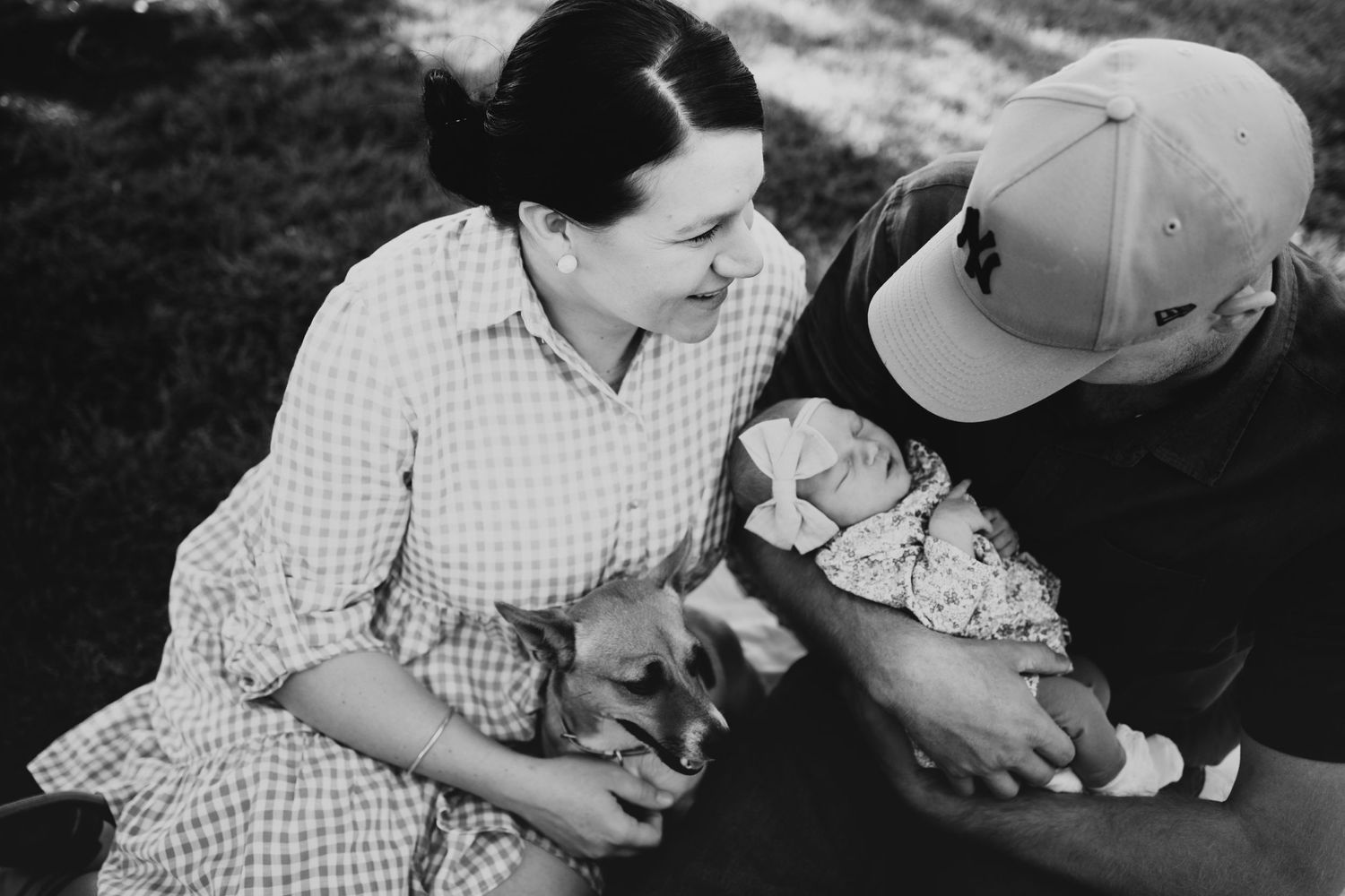 Black and white photo of a couple sitting on grass with their newborn baby and a dog.