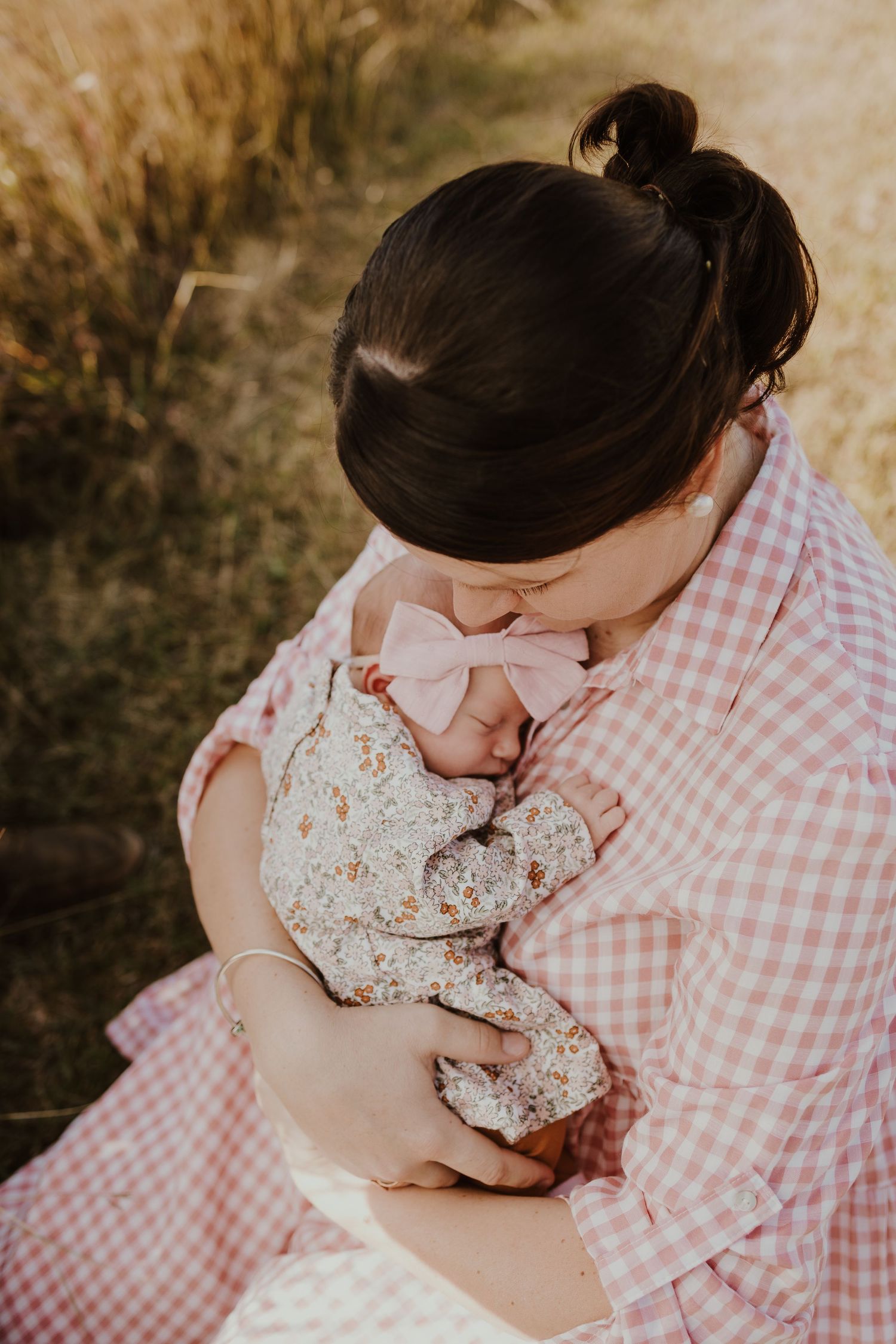 Mother in pink gingham dress tenderly holding and kissing her sleeping newborn baby girl.