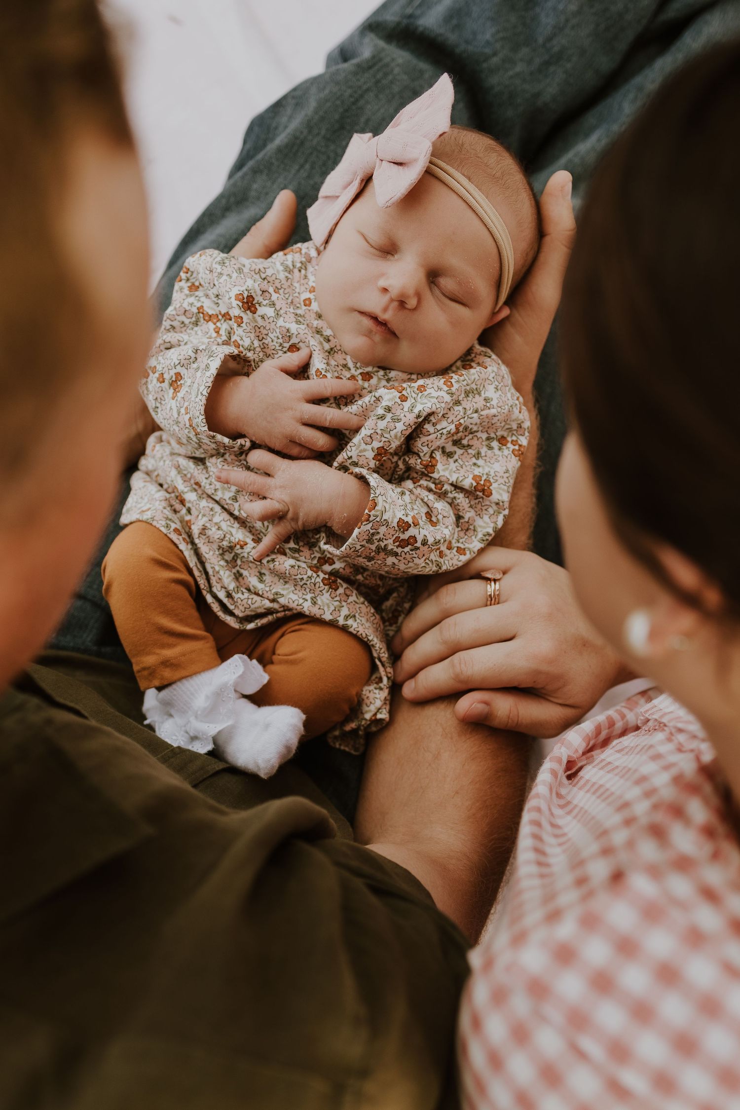 Parents hands cradling their sleeping newborn baby girl in a floral outfit and pink bow.