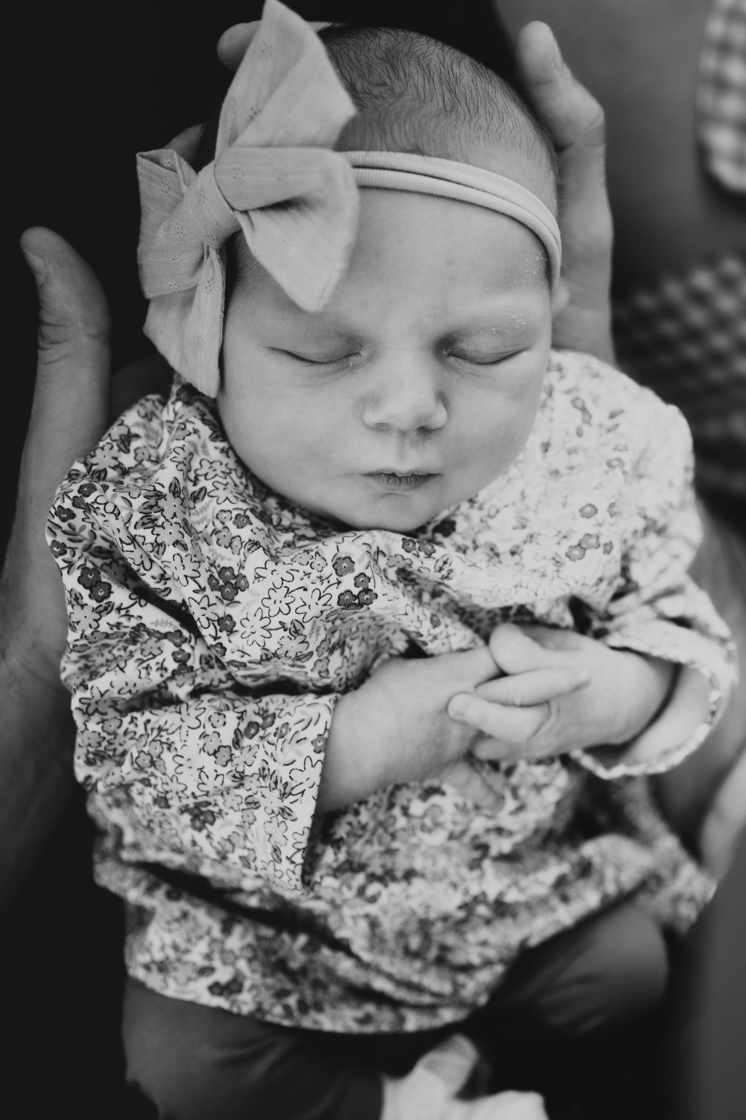 Black and white close-up of a sleeping newborn baby girl in floral outfit with pink bow.