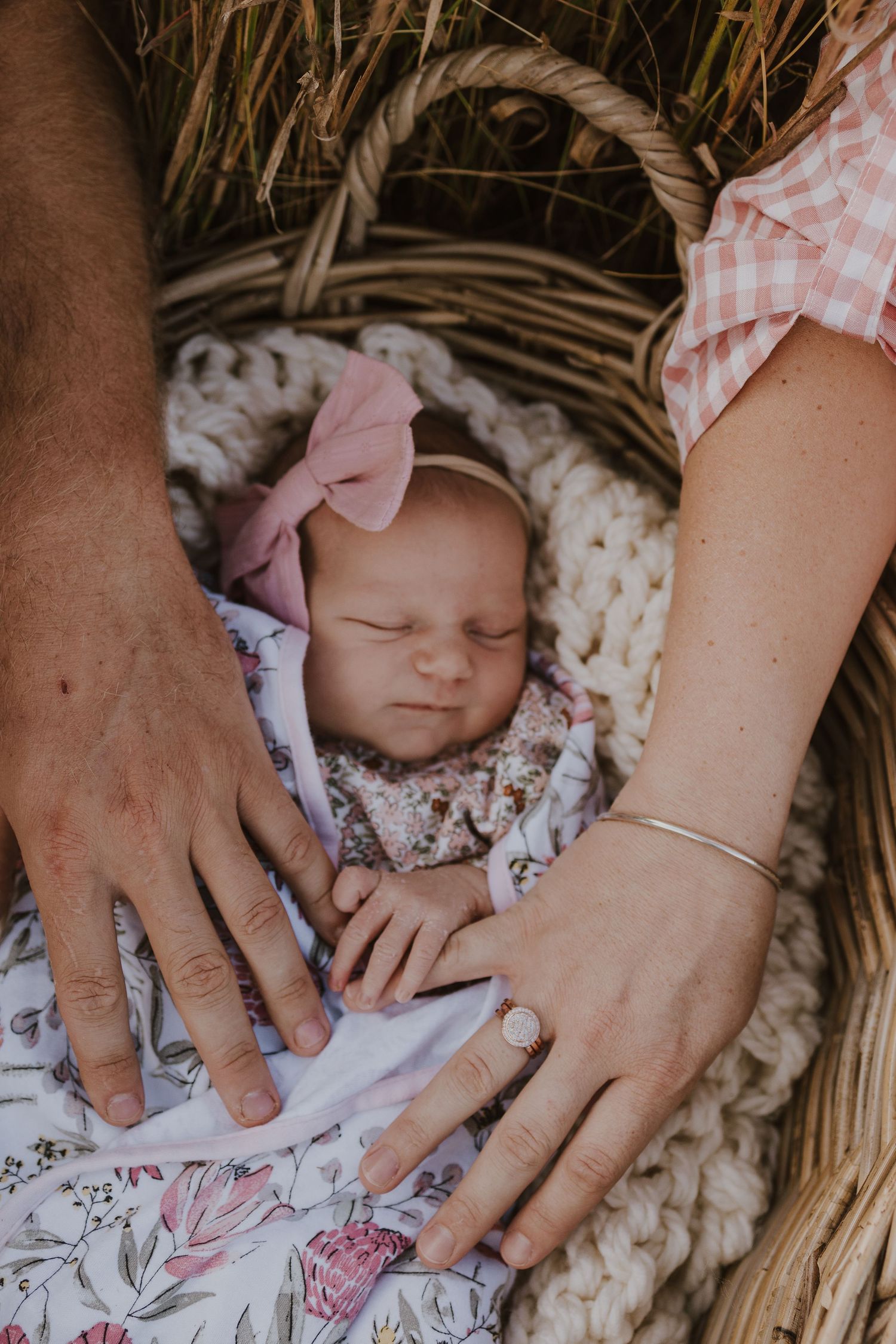 Sleeping newborn baby girl with pink bow headband resting in wicker basket with parents hands nearby.