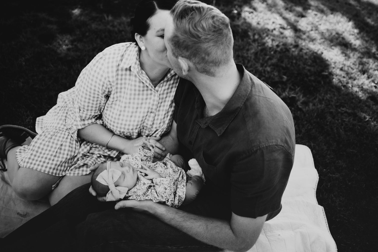 Black and white photo of a couple kissing while holding their newborn baby outdoors.