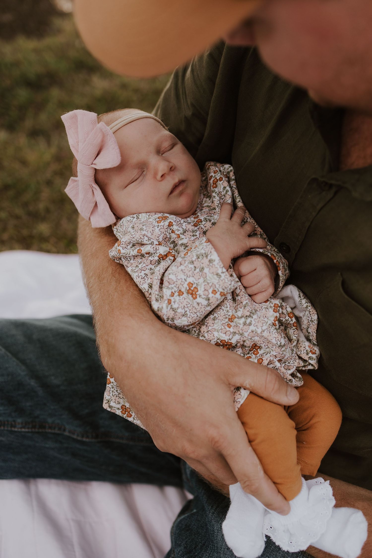 Father in green shirt gently cradling sleeping newborn baby girl with pink bow headband.
