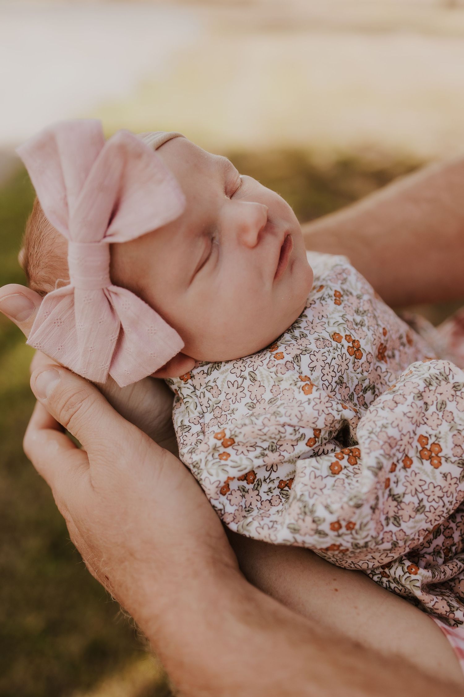 Newborn baby girl in floral outfit and pink bow headband sleeping in fathers hands.