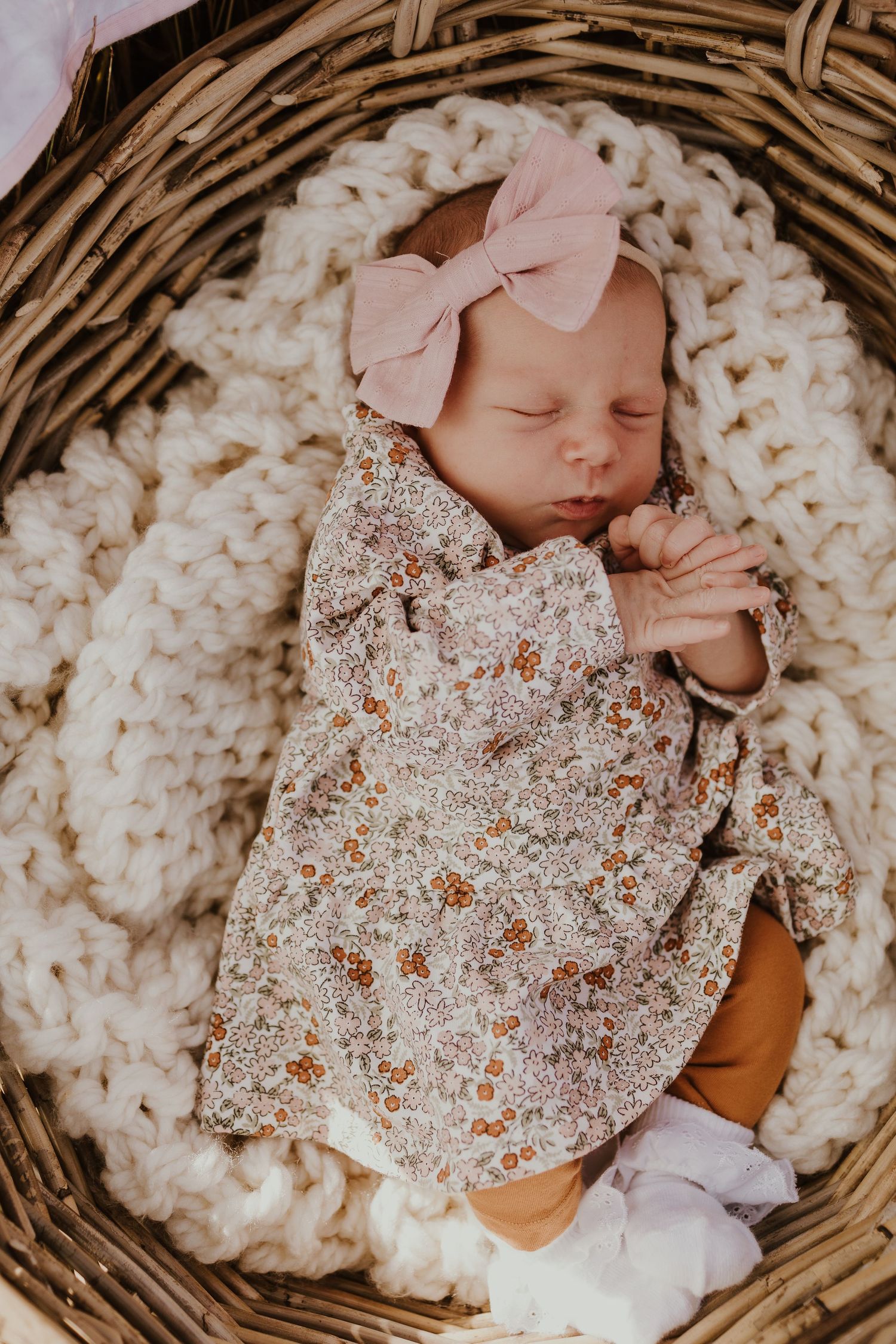 Newborn baby girl in floral outfit and pink bow headband sleeping in a wicker basket.