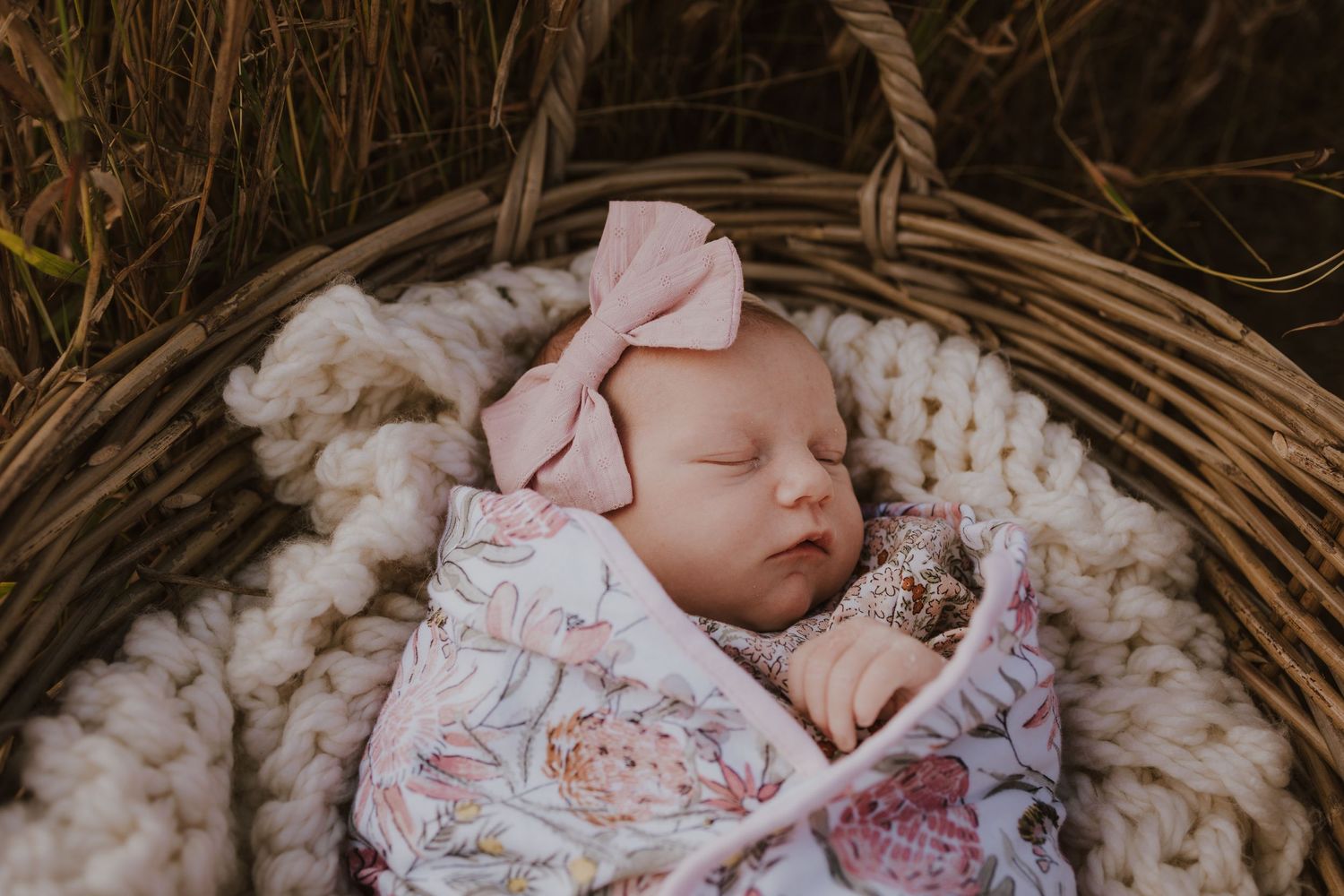 Peaceful sleeping newborn baby girl with pink bow headband swaddled in a wicker basket.