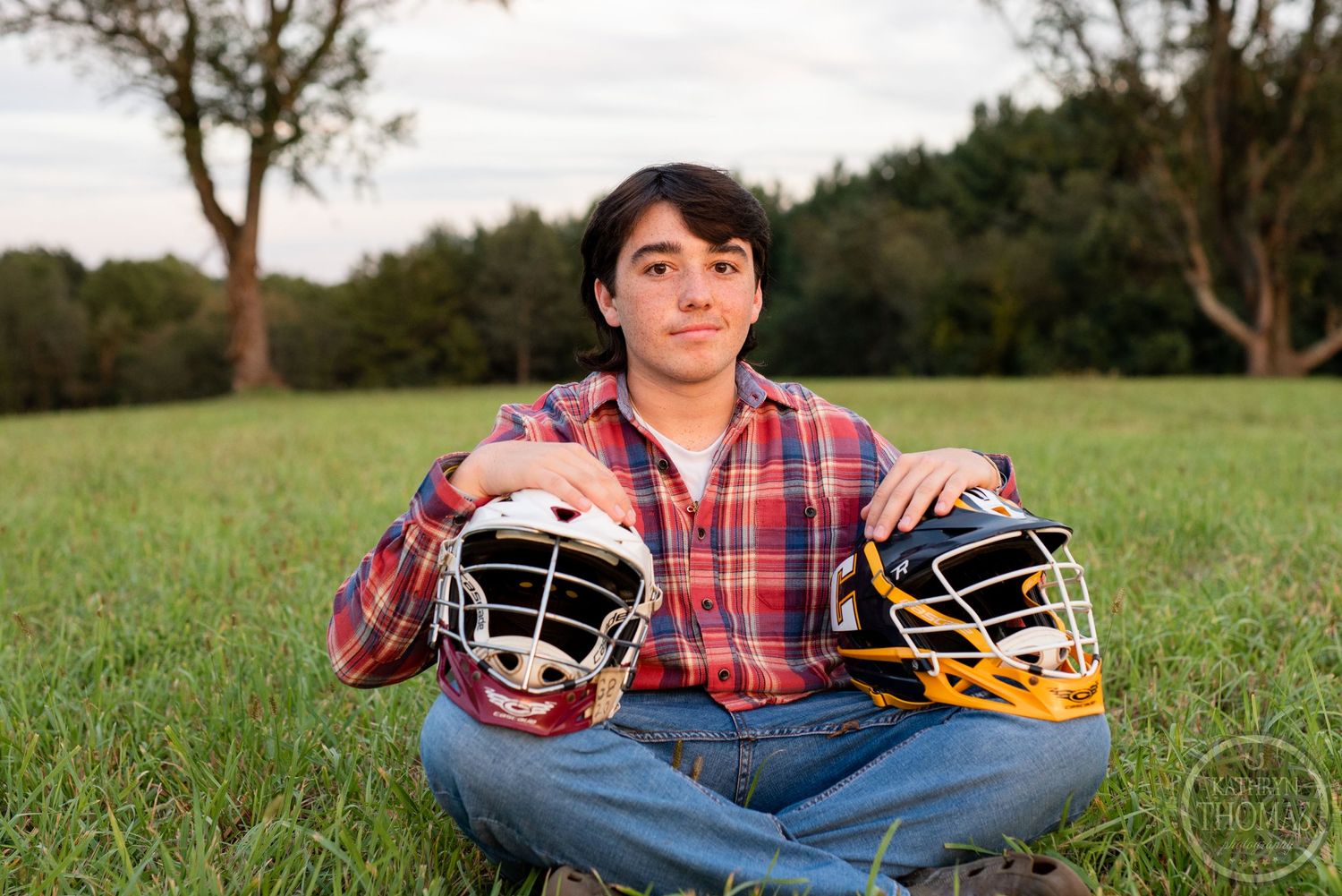 A person in a plaid shirt holds two sports helmets while sitting in a grassy field.