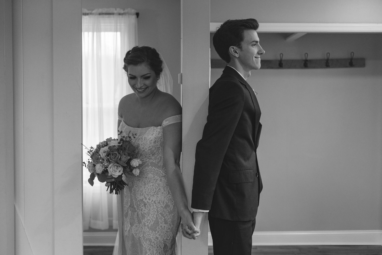 A black and white wedding photo showing a bride in a lace dress and groom in a suit standing back to back.