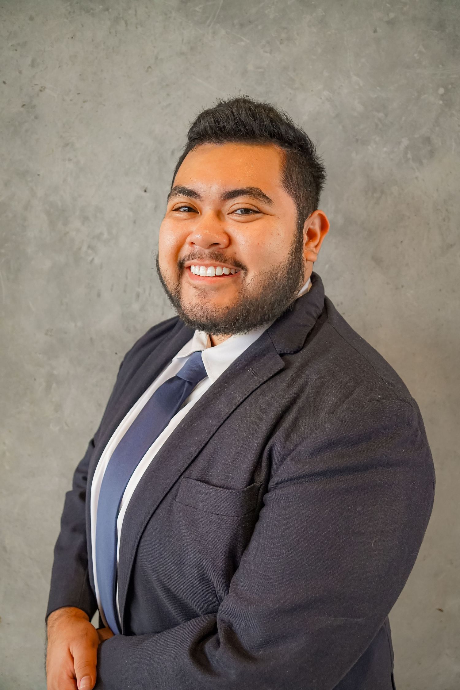 Professional headshot of person in dark suit and tie against concrete wall.