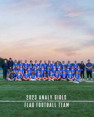 Large group photo of girls flag football team in blue uniforms on sports field at sunset.