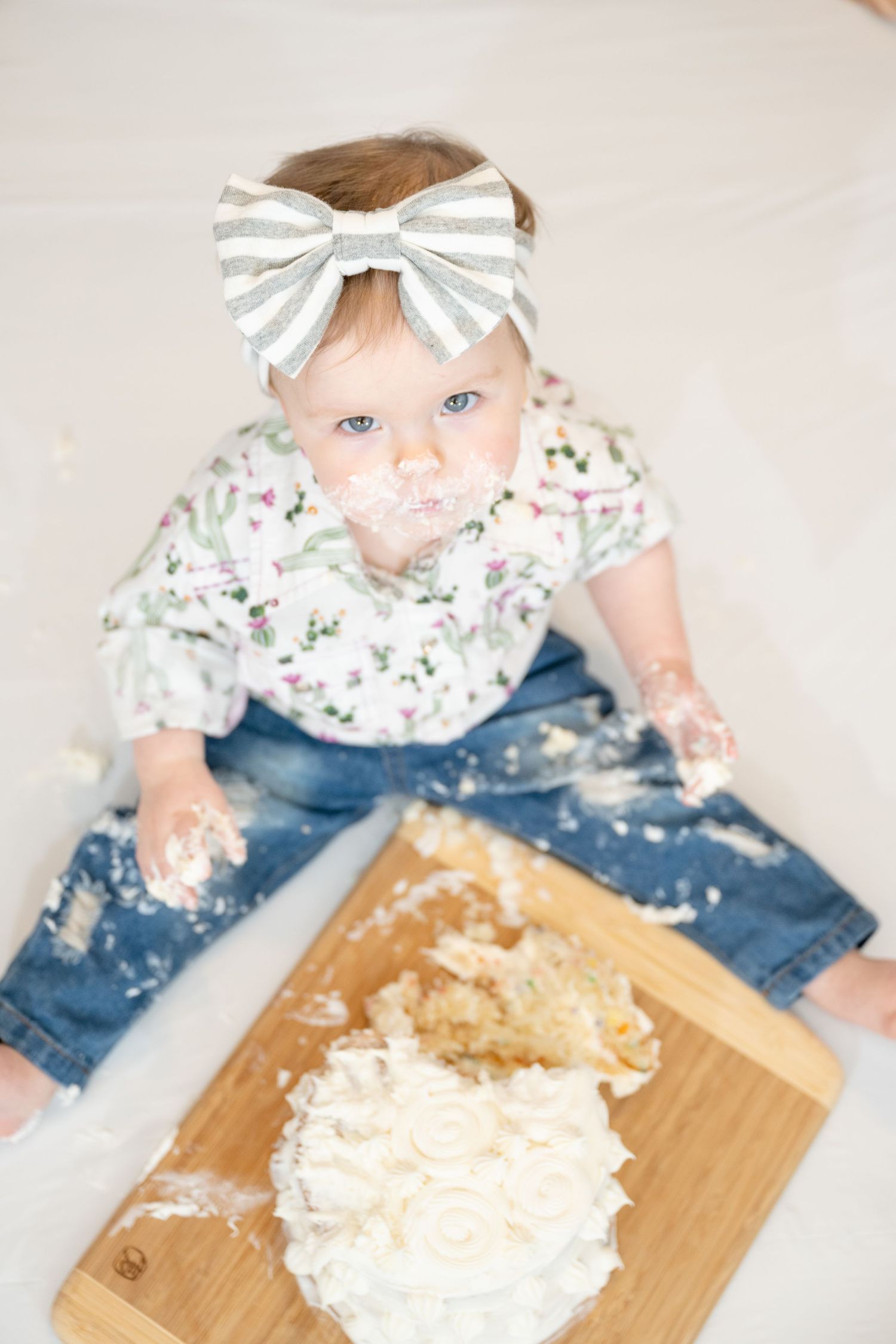 A baby in floral outfit and striped bow plays with cake on wooden board.