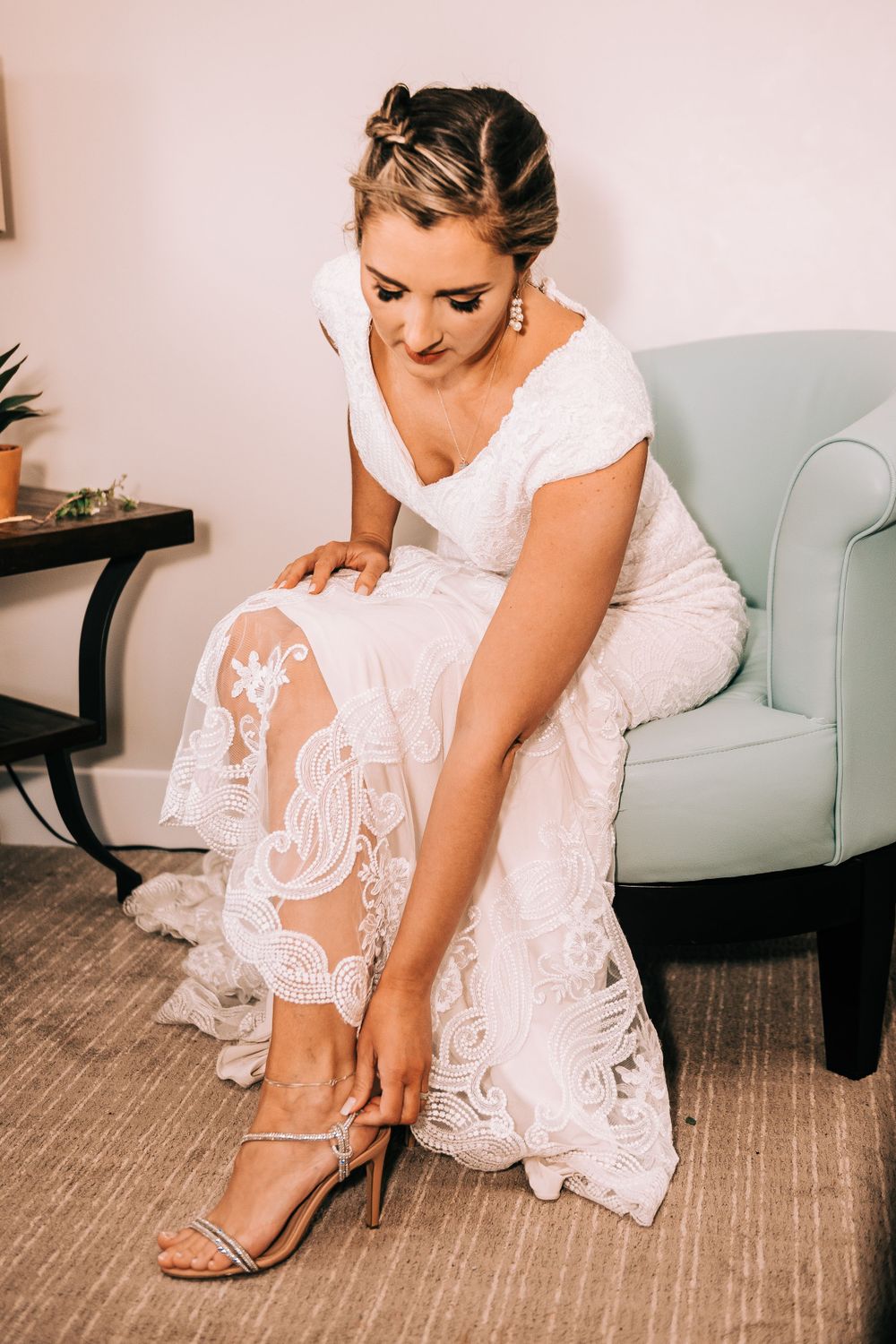 Person in white lace wedding dress adjusting strappy sandal while seated on blue chair.