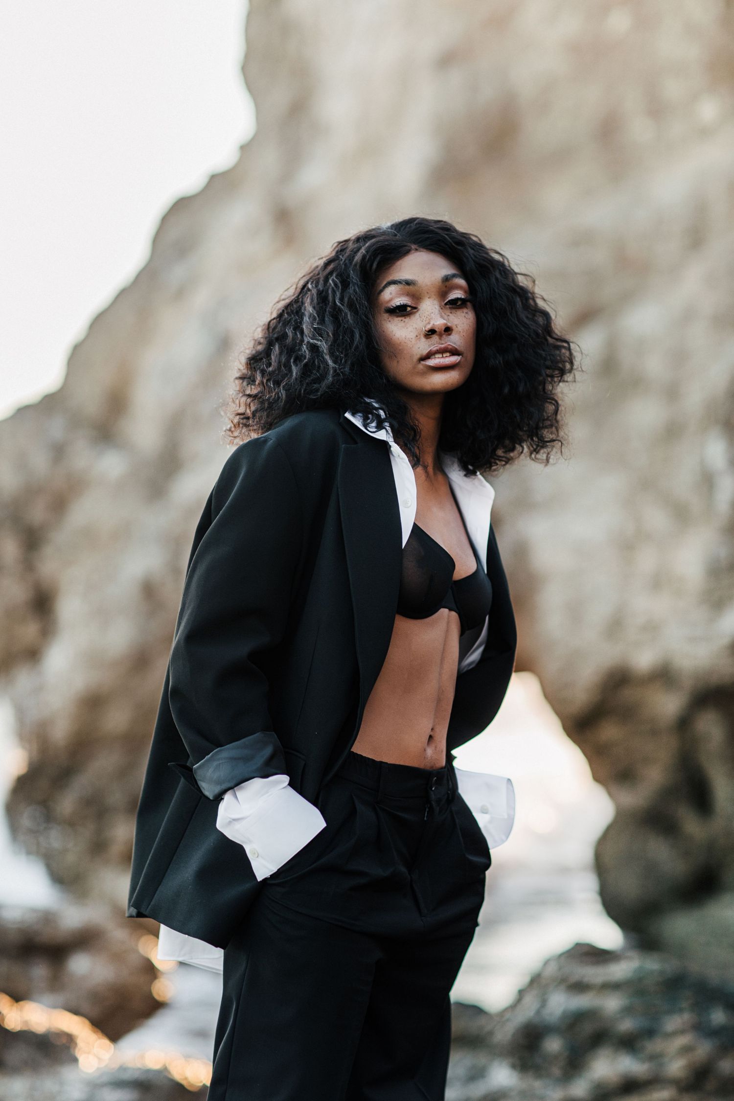 Professional fashion model poses on rocky beach wearing black blazer and bralette with natural curly hair.