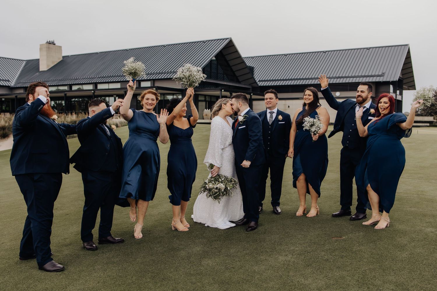 Wedding party dances and celebrates on a grassy venue with rustic buildings in the background.