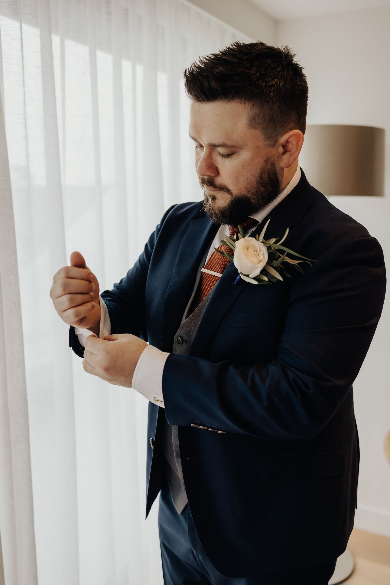 Groom adjusts tie while getting ready in bright white room.