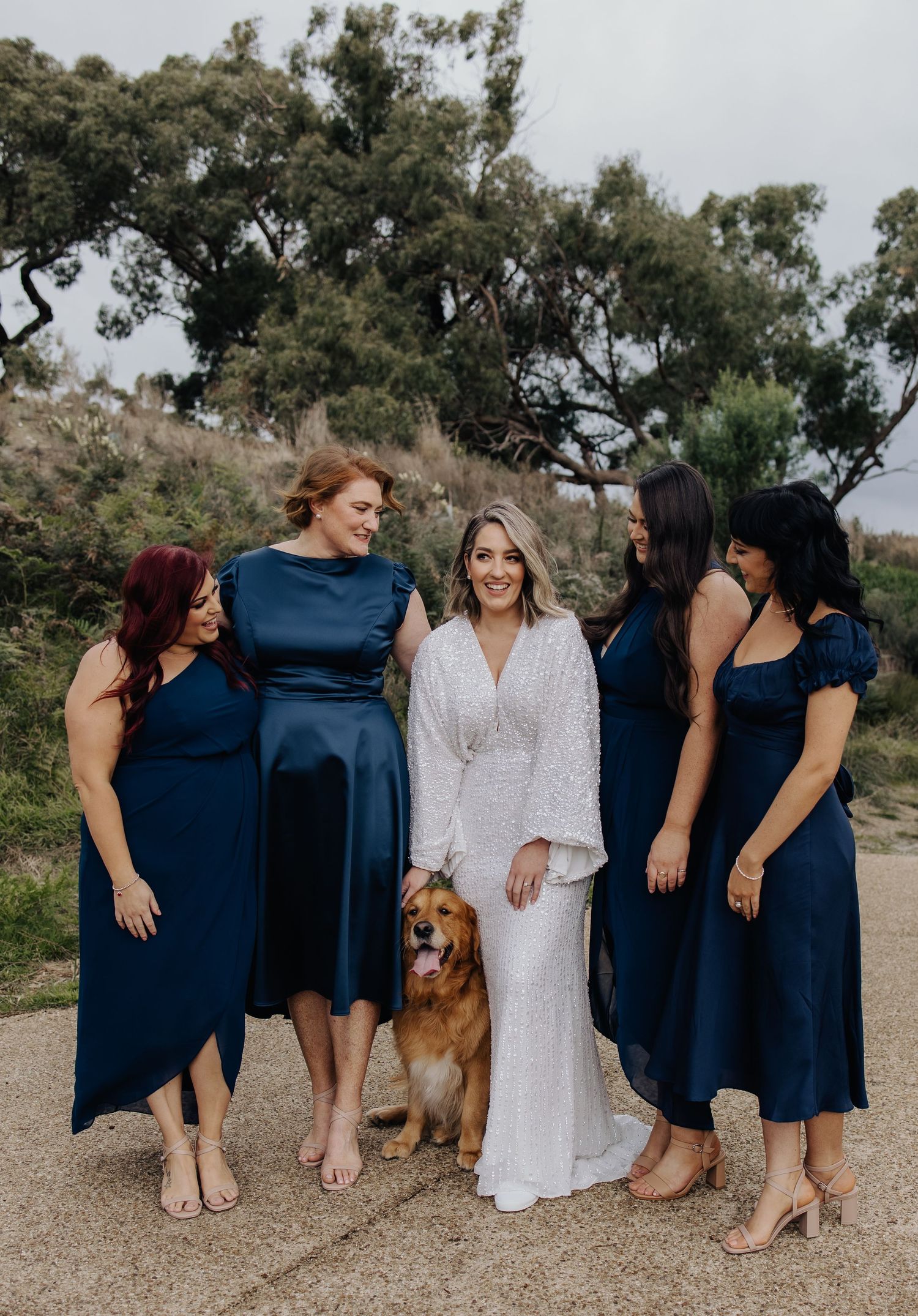 Bridesmaids in navy dresses and a bride in white pose with a friendly golden retriever at an outdoor wedding.