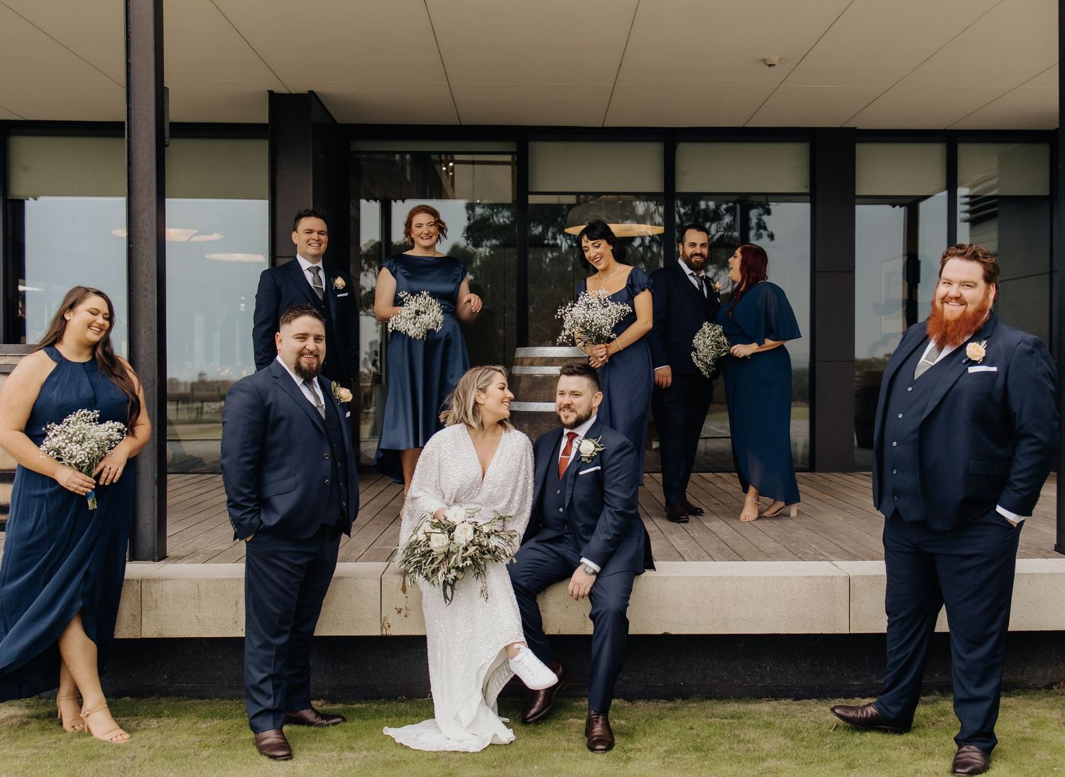 Wedding group sitting on ground against modern building exterior.