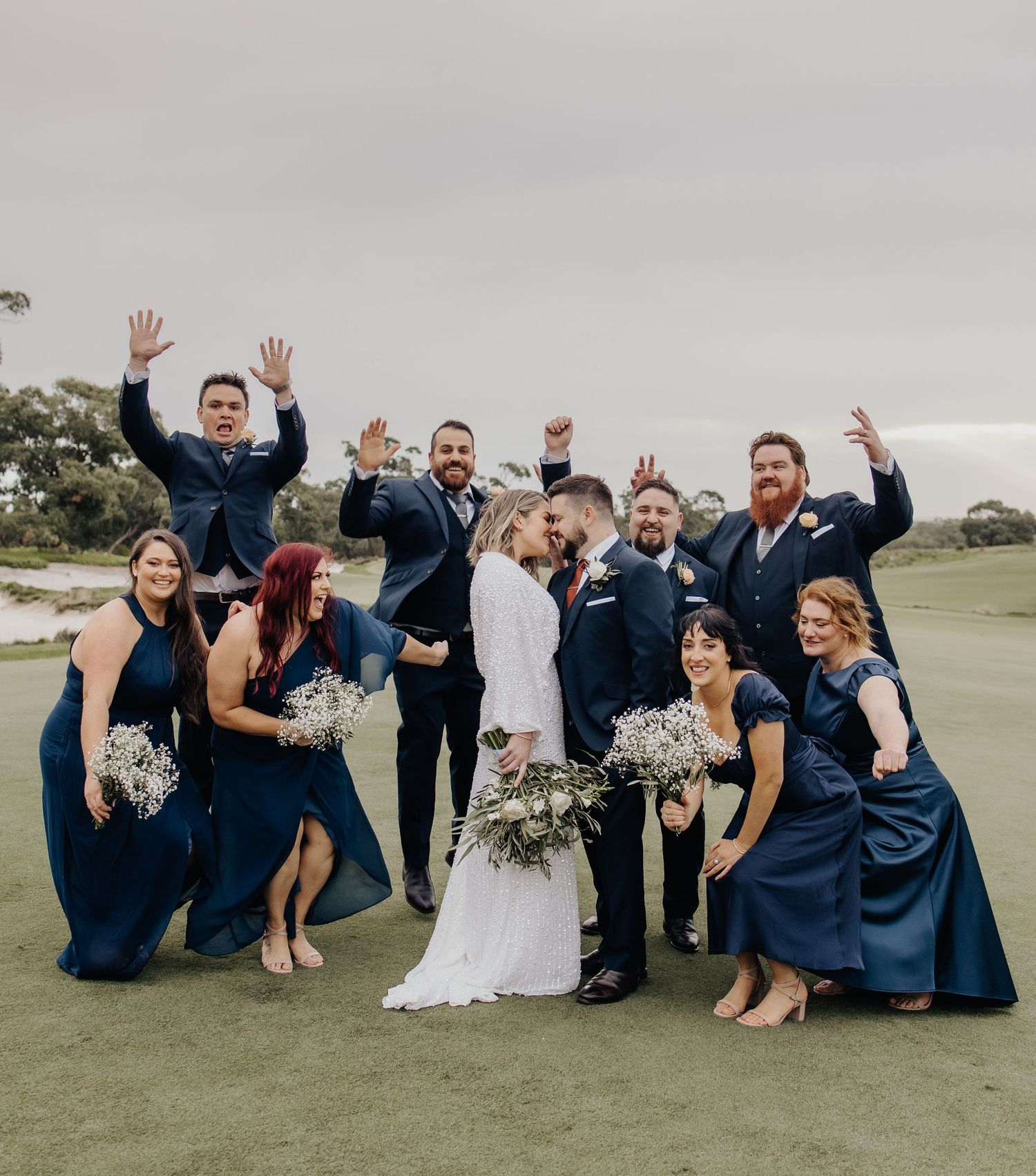 Wedding party poses playfully on grass in navy blue and white attire with bouquets.