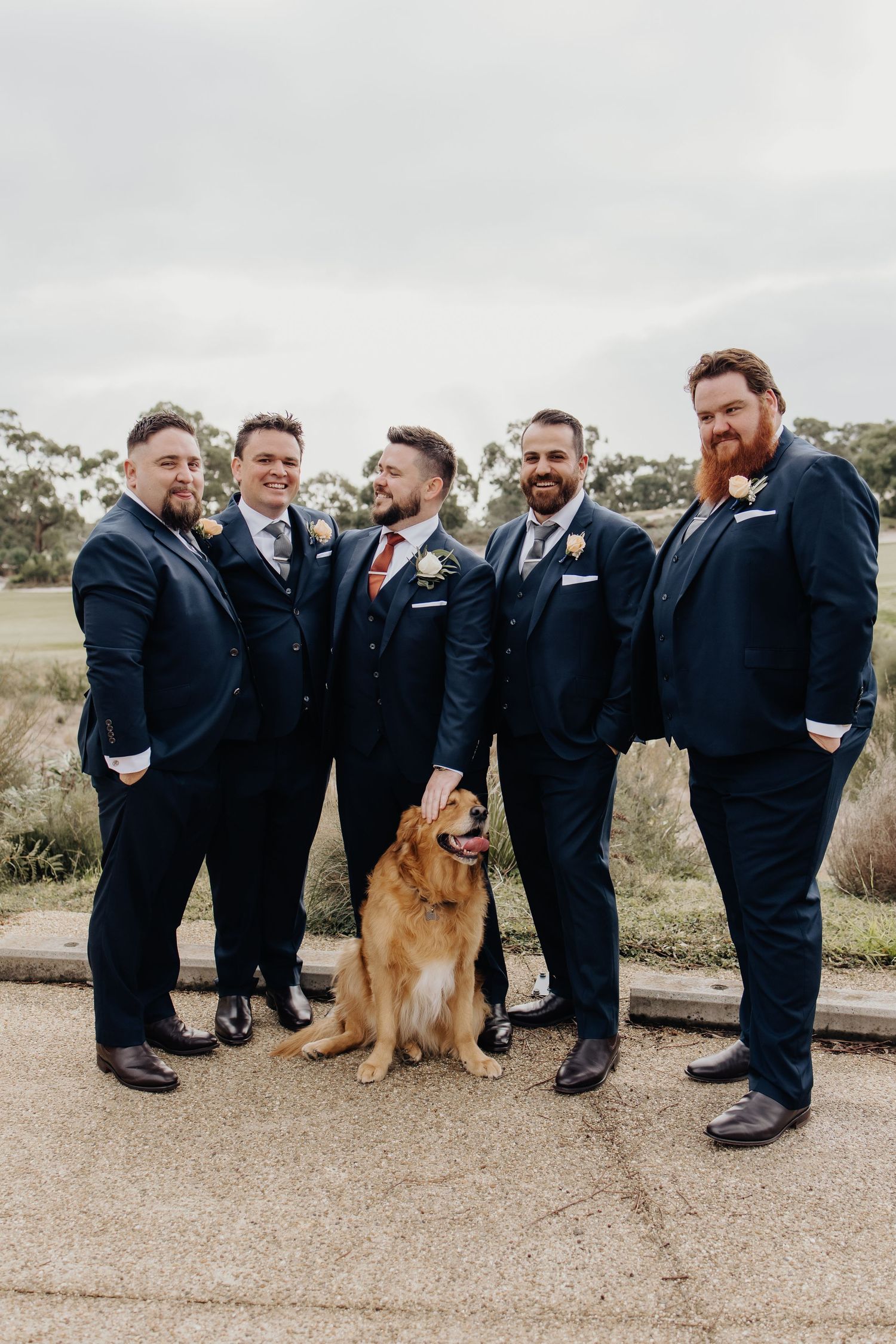 Groomsmen in navy suits pose playfully with a golden retriever on a gravel path during a wedding photo shoot.