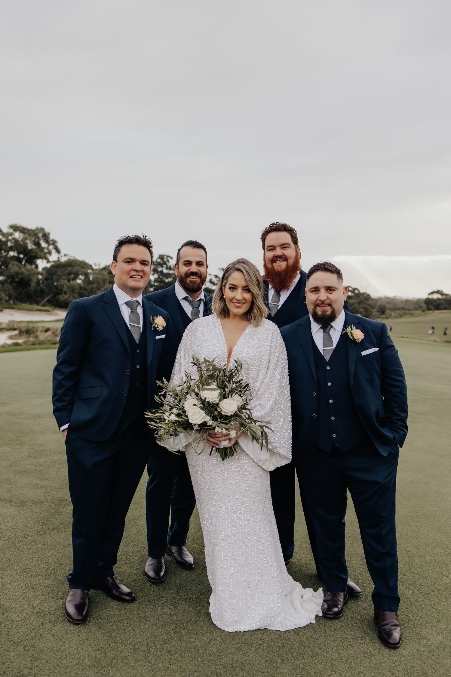 Wedding party members pose together on a green lawn during an overcast day.