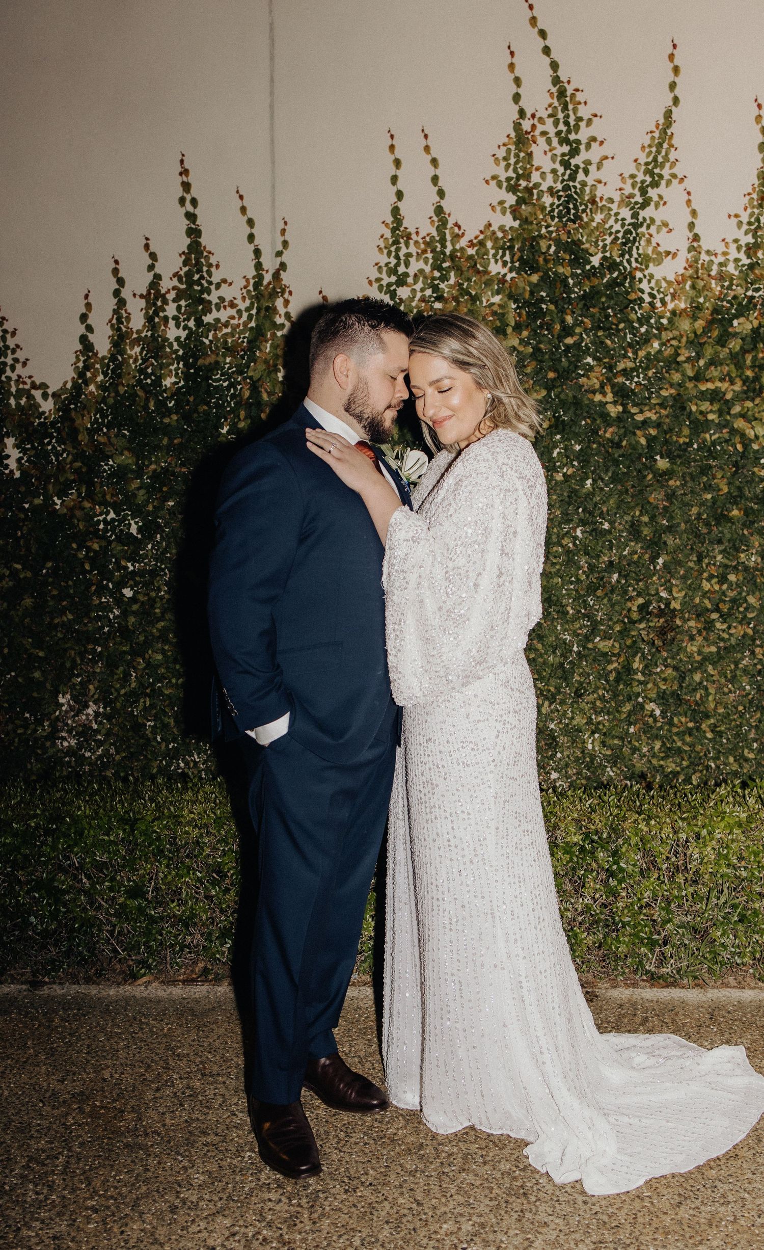 Newlyweds share intimate moment against garden greenery at night.
