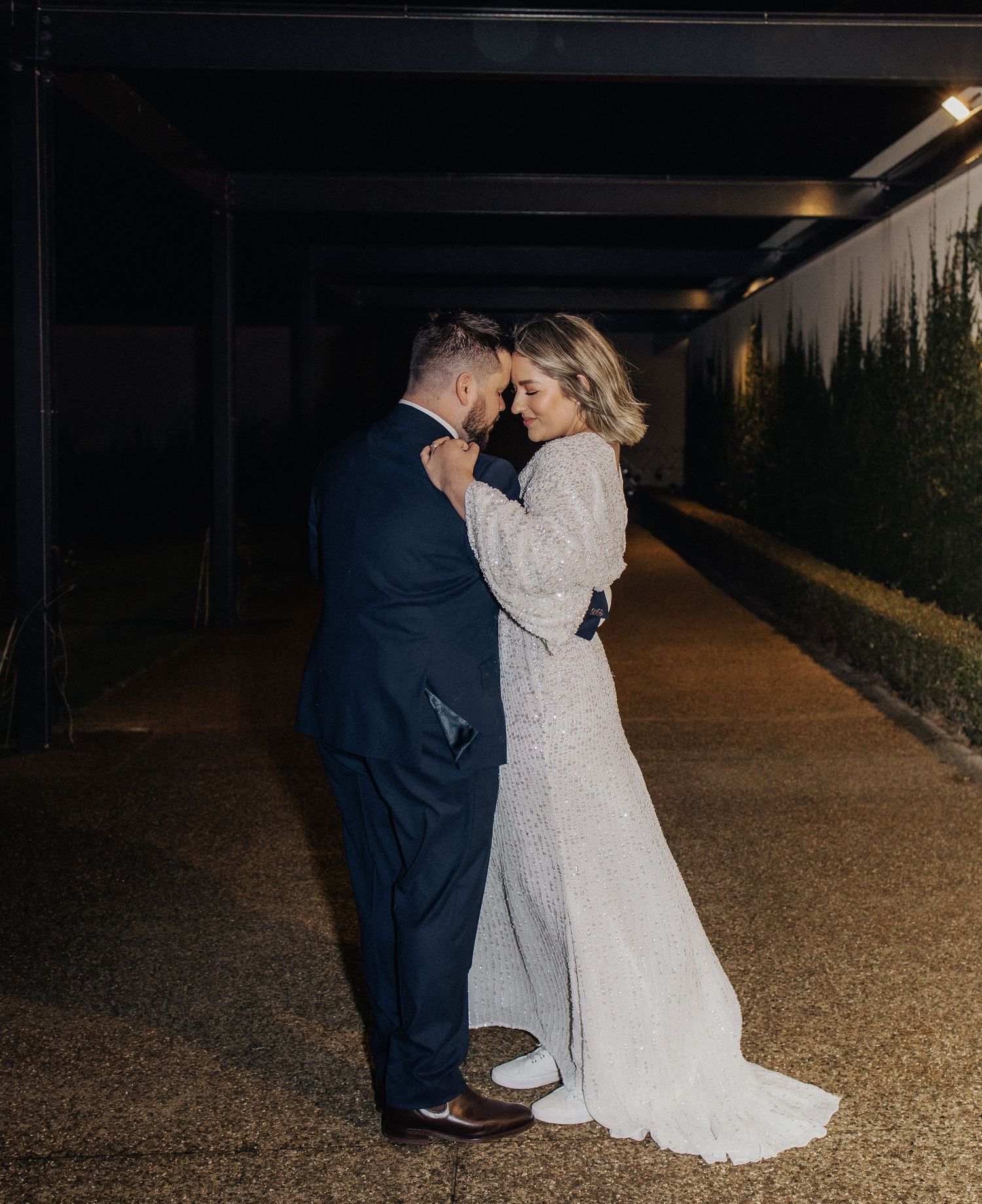 Bride and groom embrace on dimly lit pathway during evening reception.