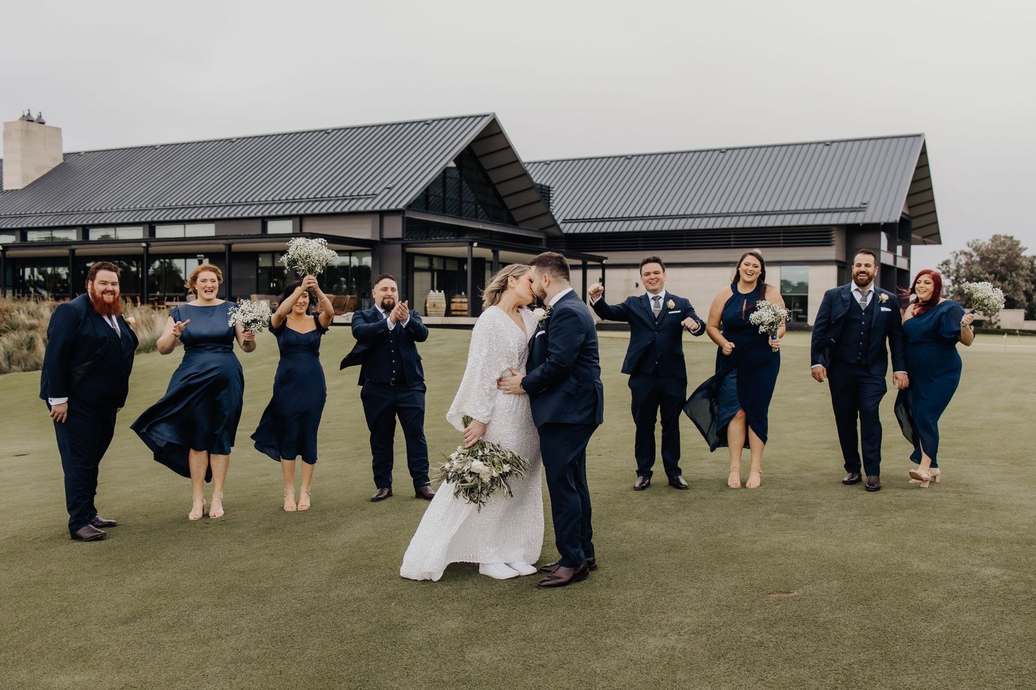 Wedding celebration with guests dancing and celebrating on a lawn near grey-roofed buildings.