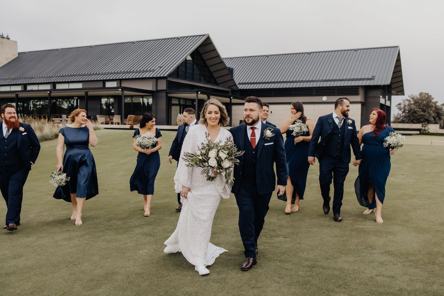 Wedding party members walk across a green lawn with rustic buildings in the background.
