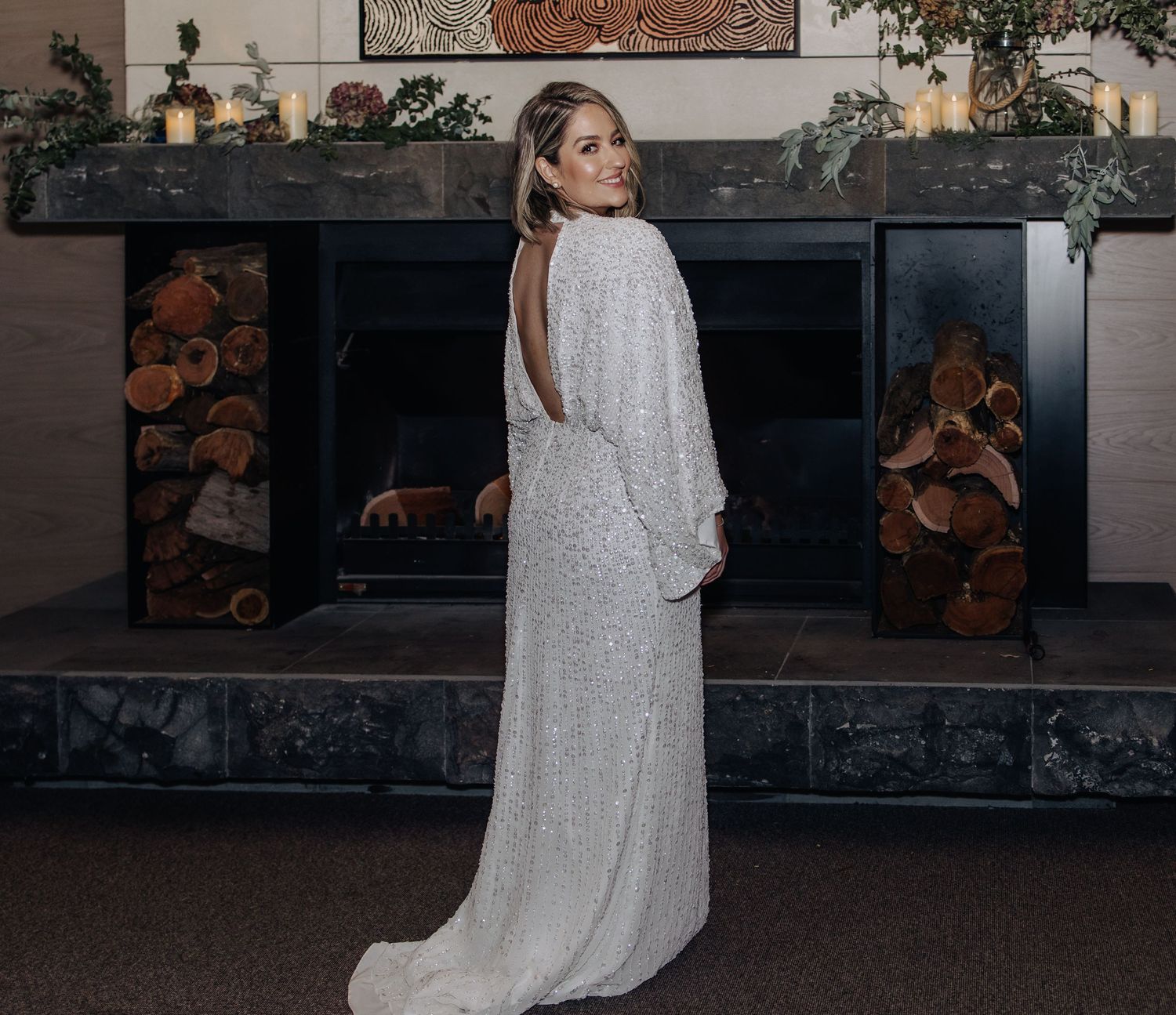 Bride in sequined gown poses near rustic fireplace with stacked wood.