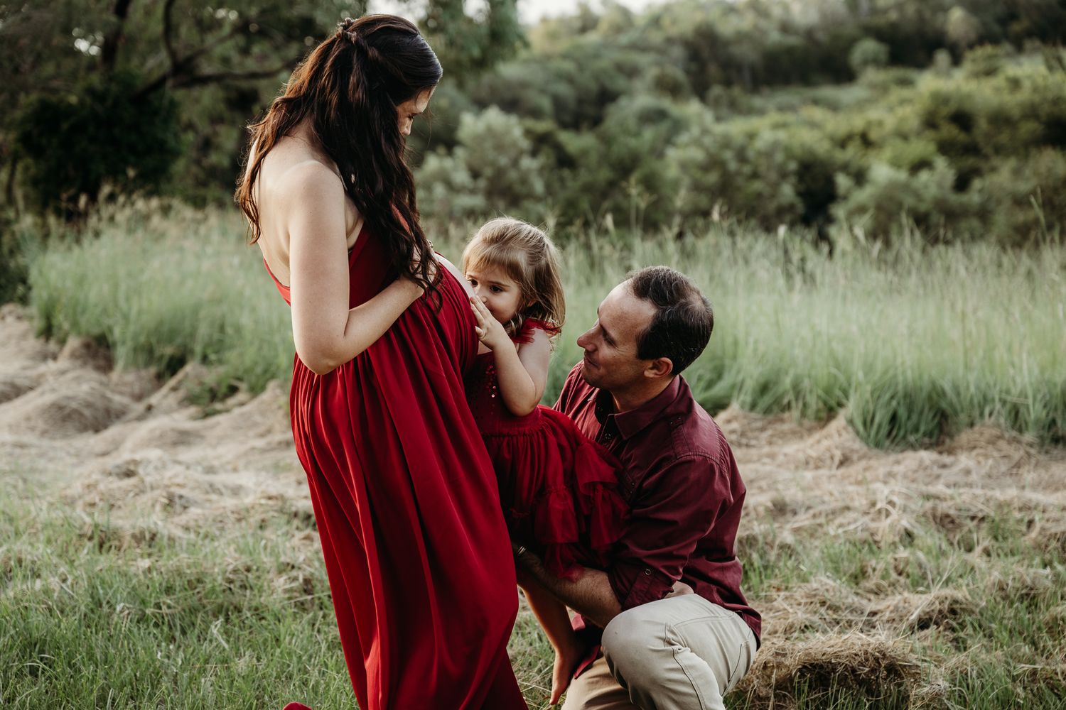 Family maternity photo in a field with people wearing red and burgundy outfits.