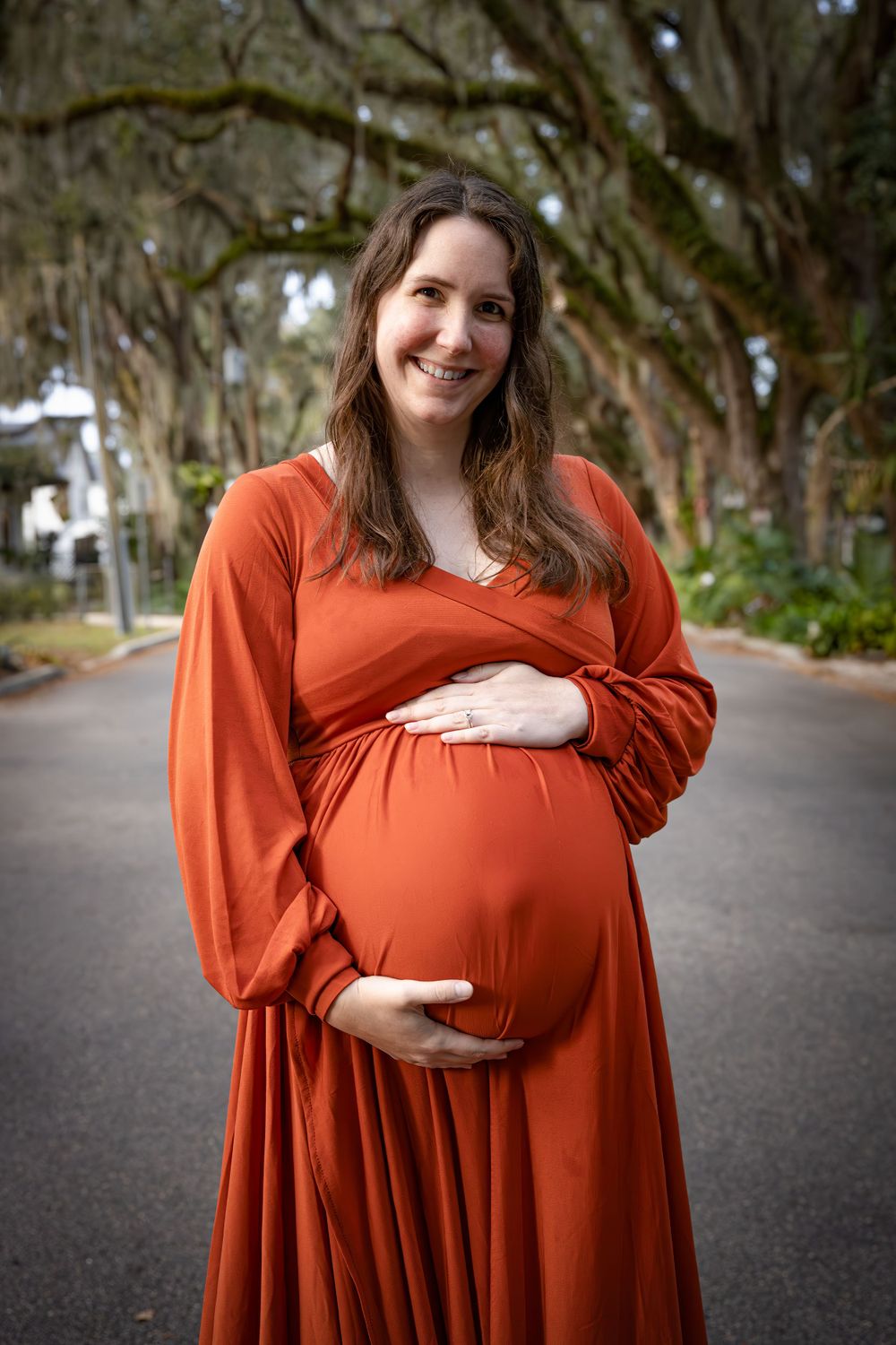 A woman in a flowing rust-colored dress stands on a tree-lined path during a maternity photoshoot.