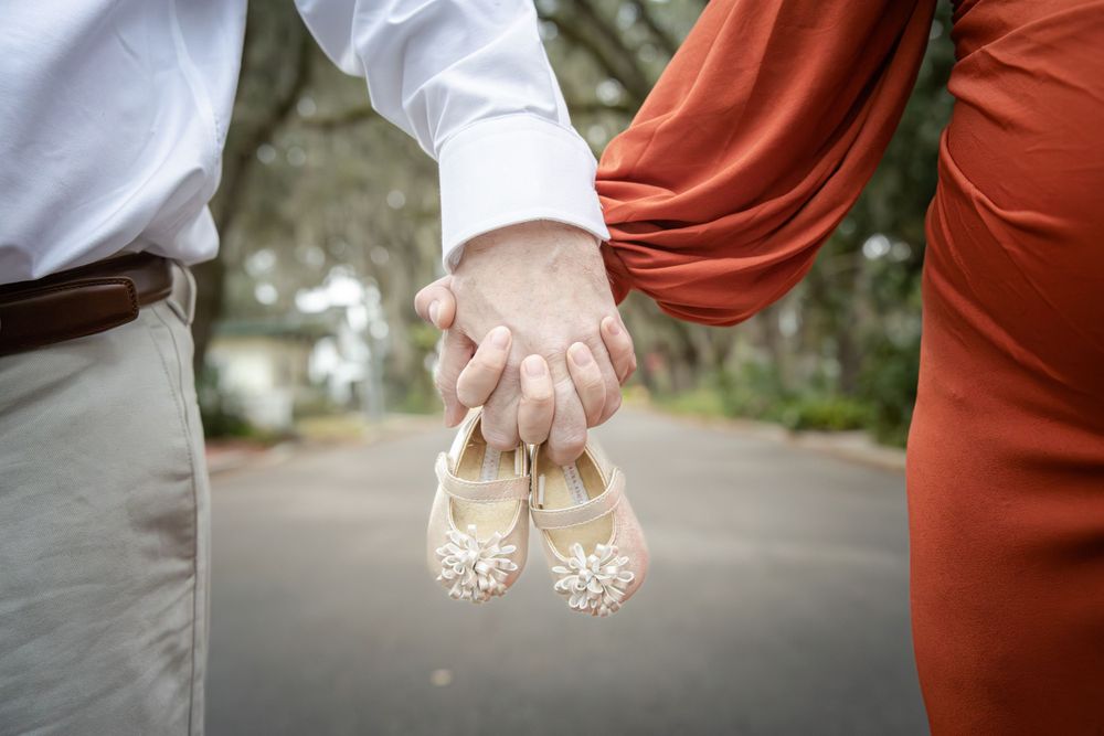 Hands holding delicate beige wedding shoes on a paved pathway.