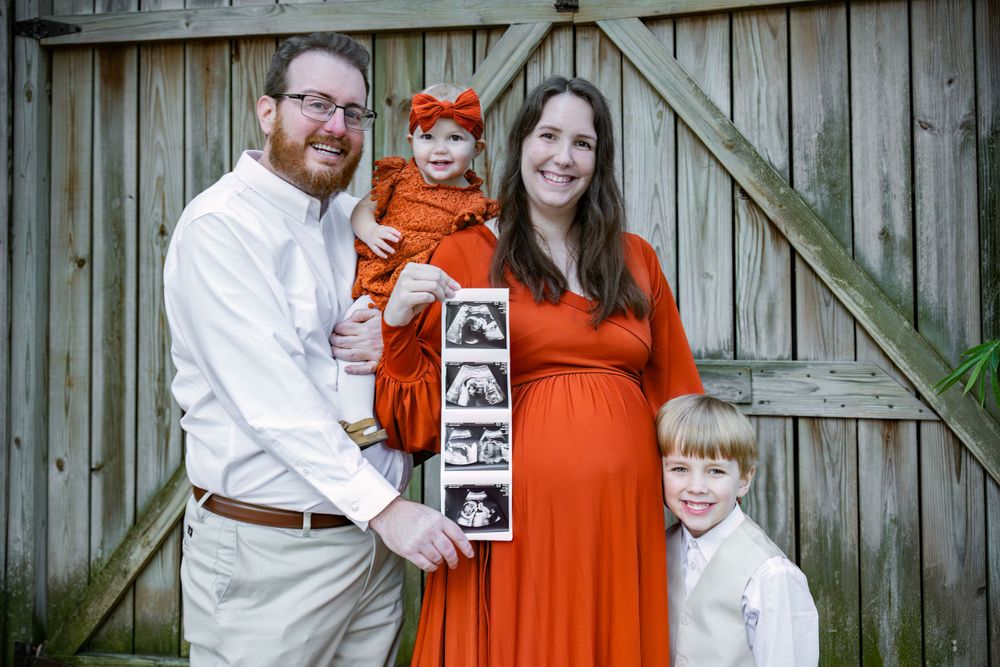 A family poses together against a wooden fence holding ultrasound photos during a pregnancy announcement.