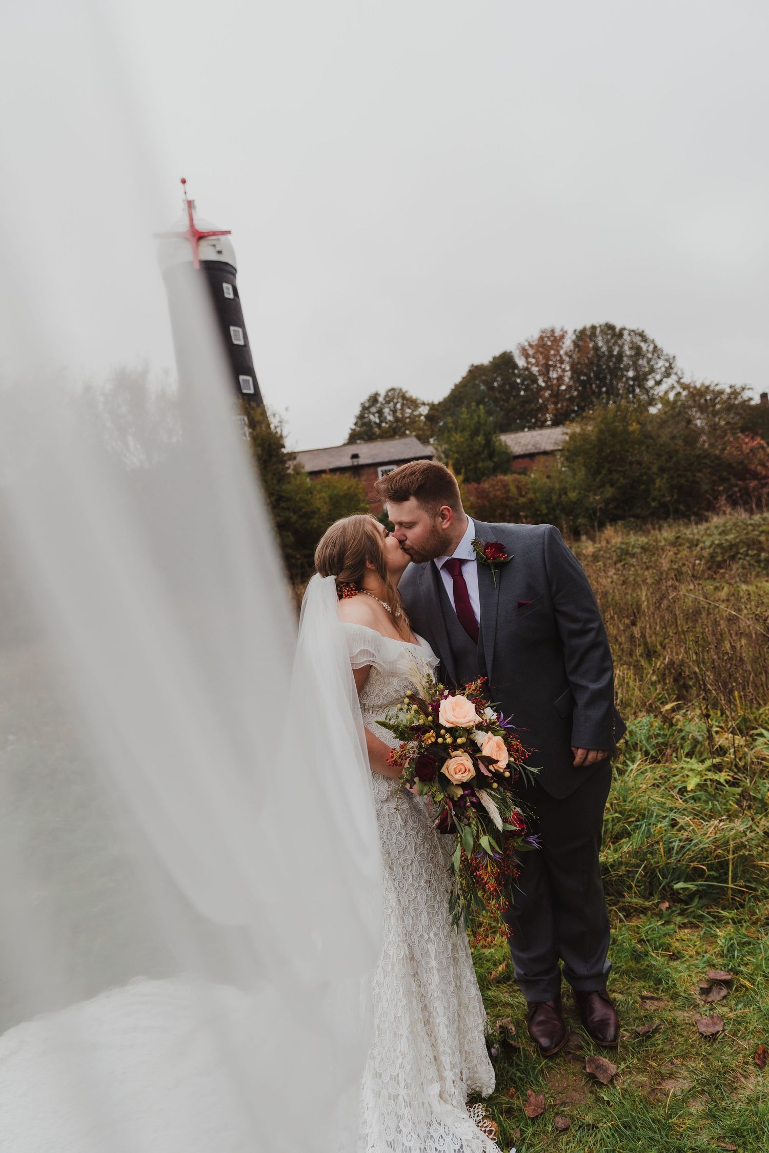 Romantic wedding couple embracing outdoors near lighthouse on overcast day.