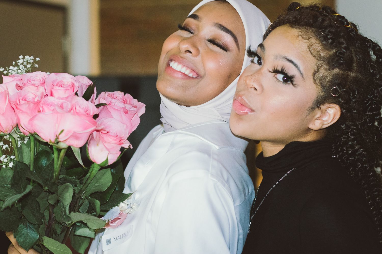 a hijabi woman smiling and holding a pink flower bouquet in front of another woman of color