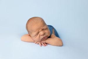 Sleeping newborn resting head on crossed arms against soft blue background.