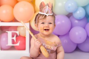 Baby in pink crown posing against rainbow balloon backdrop for first birthday celebration.