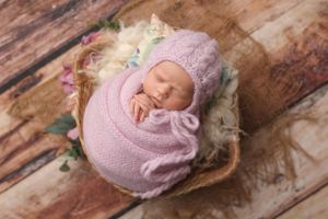 Sleeping newborn wrapped in pink knit swaddle and hat rests in rustic wooden bowl with floral accents.