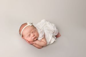 Sleeping newborn wearing lace dress and floral headband on light backdrop.
