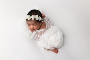 Newborn in white dress and floral crown sleeps peacefully against white background.