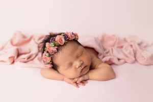 Sleeping newborn wearing delicate pink floral crown on soft pink backdrop.