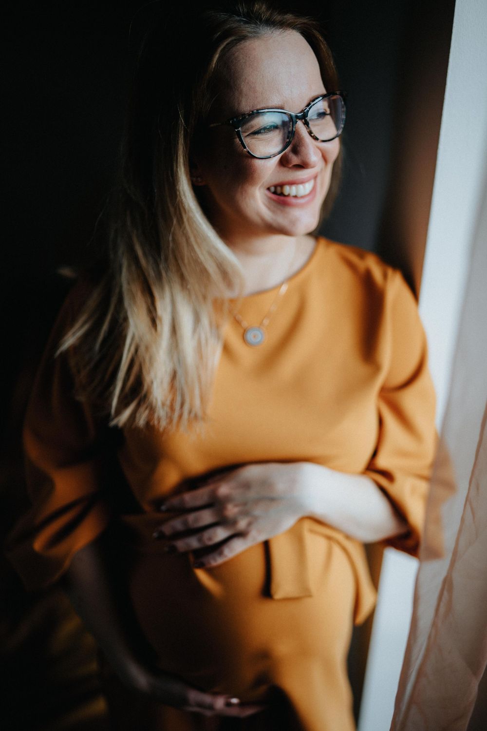A pregnant woman in an orange sweater smiles by a window.